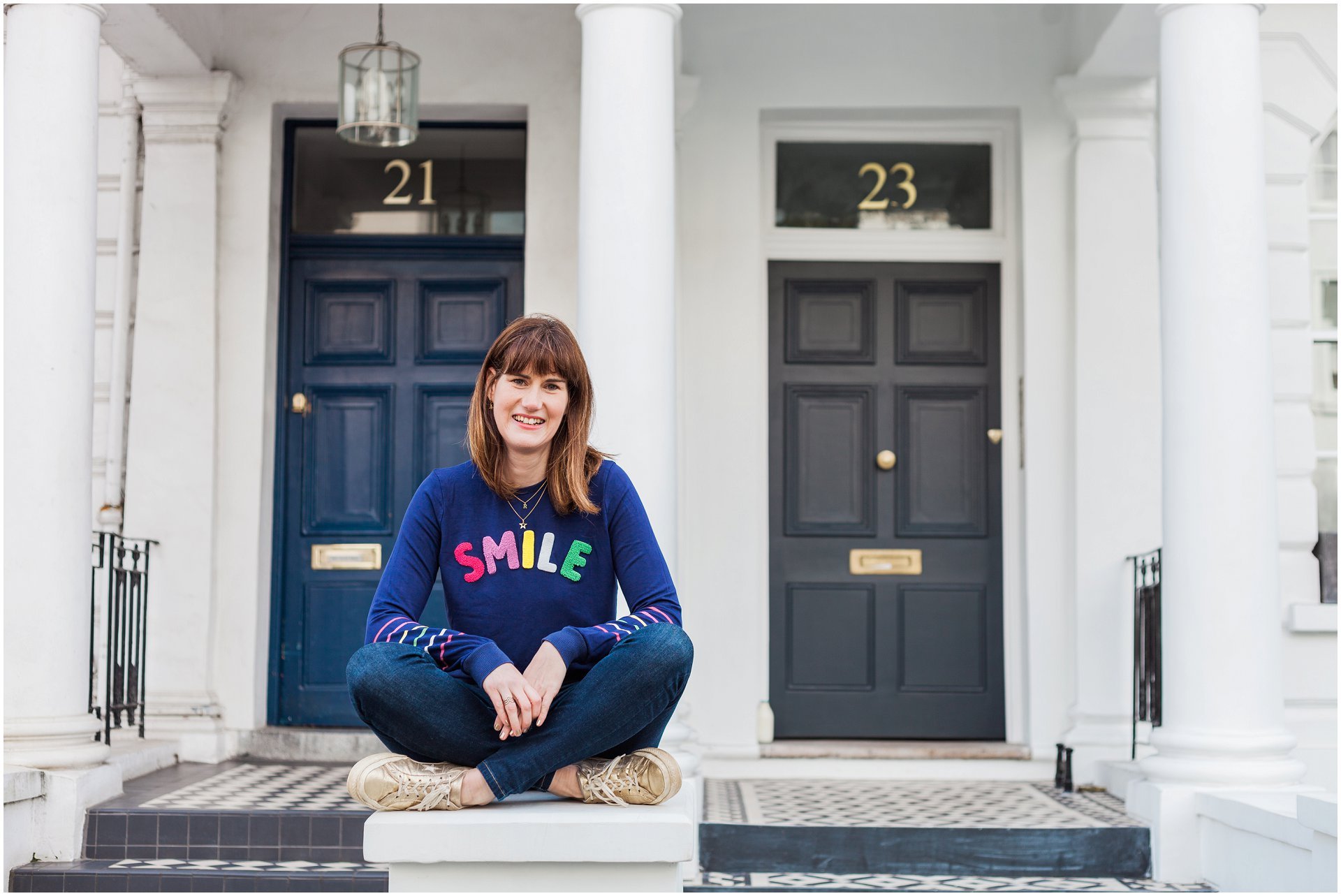 FB Ads coach Becky Wise sitting on a wall in Notting Hill wearing a blue smile jumper in her London brand shoot. Image by London branding photographer AKP Branding Stories