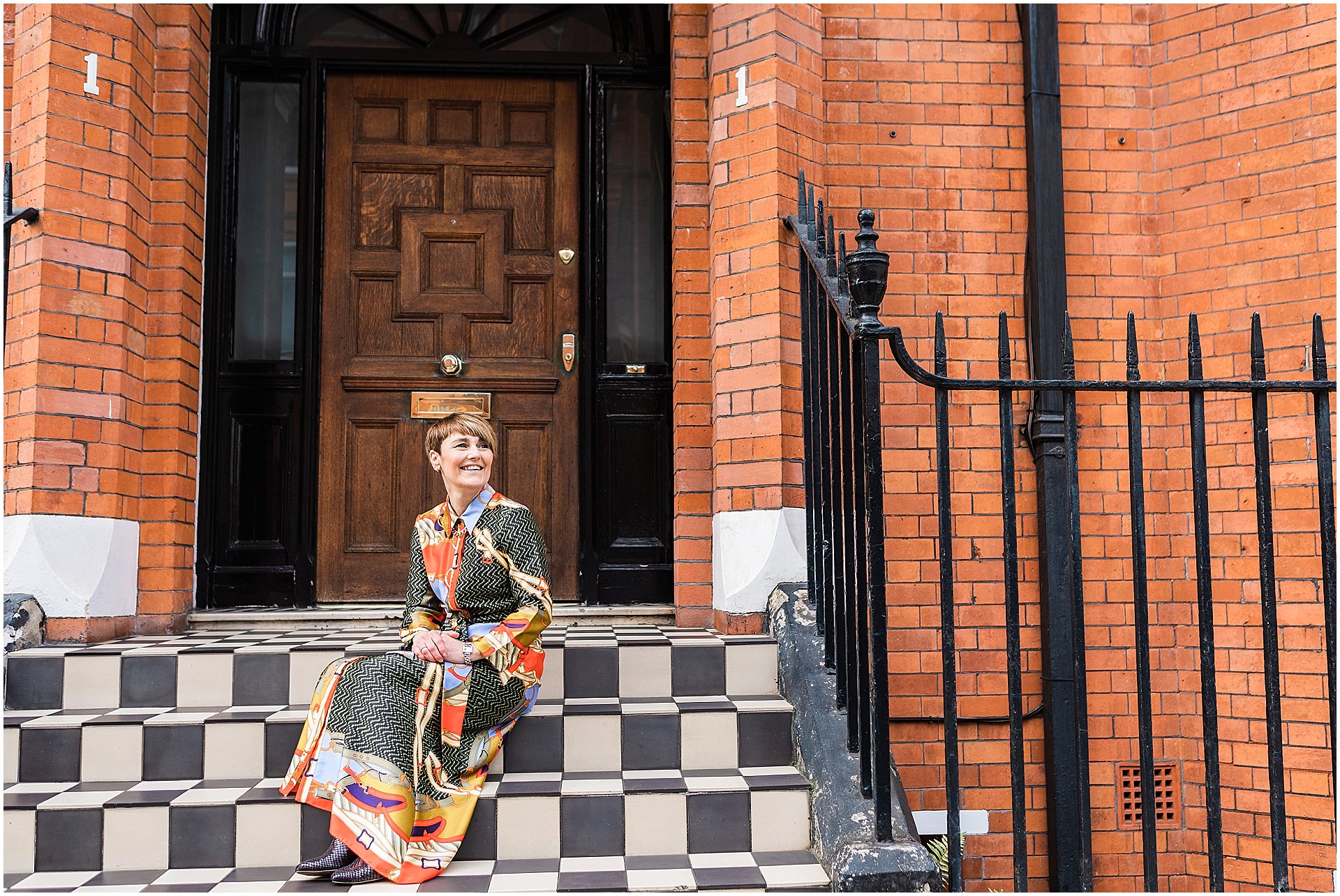 Lisa Talbot, stylist is sitting on some chequered steps in front of a red brick building. She is a wearing a patterned dress. Images by London brand photographer AKP Branding Stories