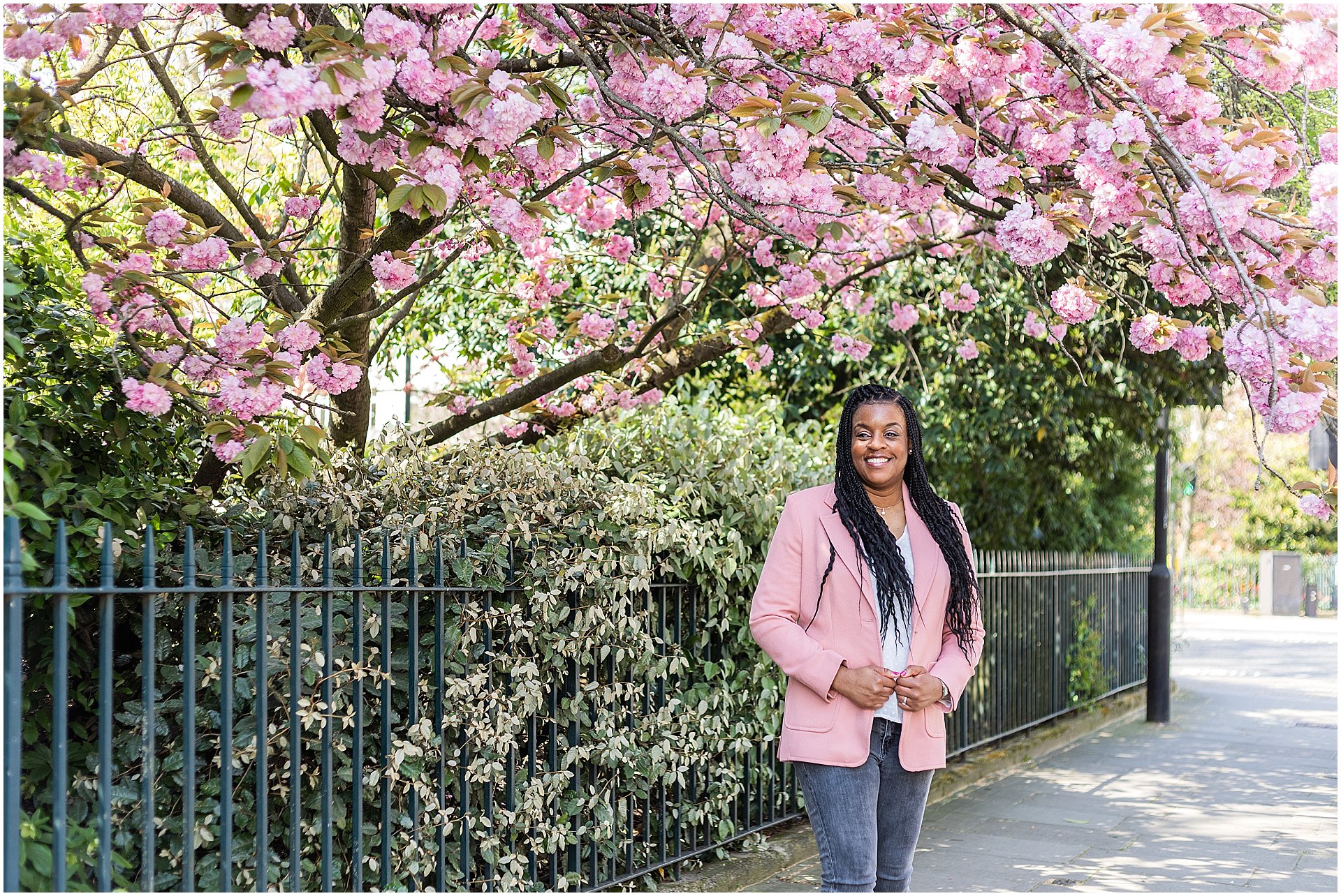 Wedding planner Natasha is stood underneath a blossom tree in London.  She is a black lady wearing jeans and a pink blazer. Image by London brand photographer AKP Branding Stories