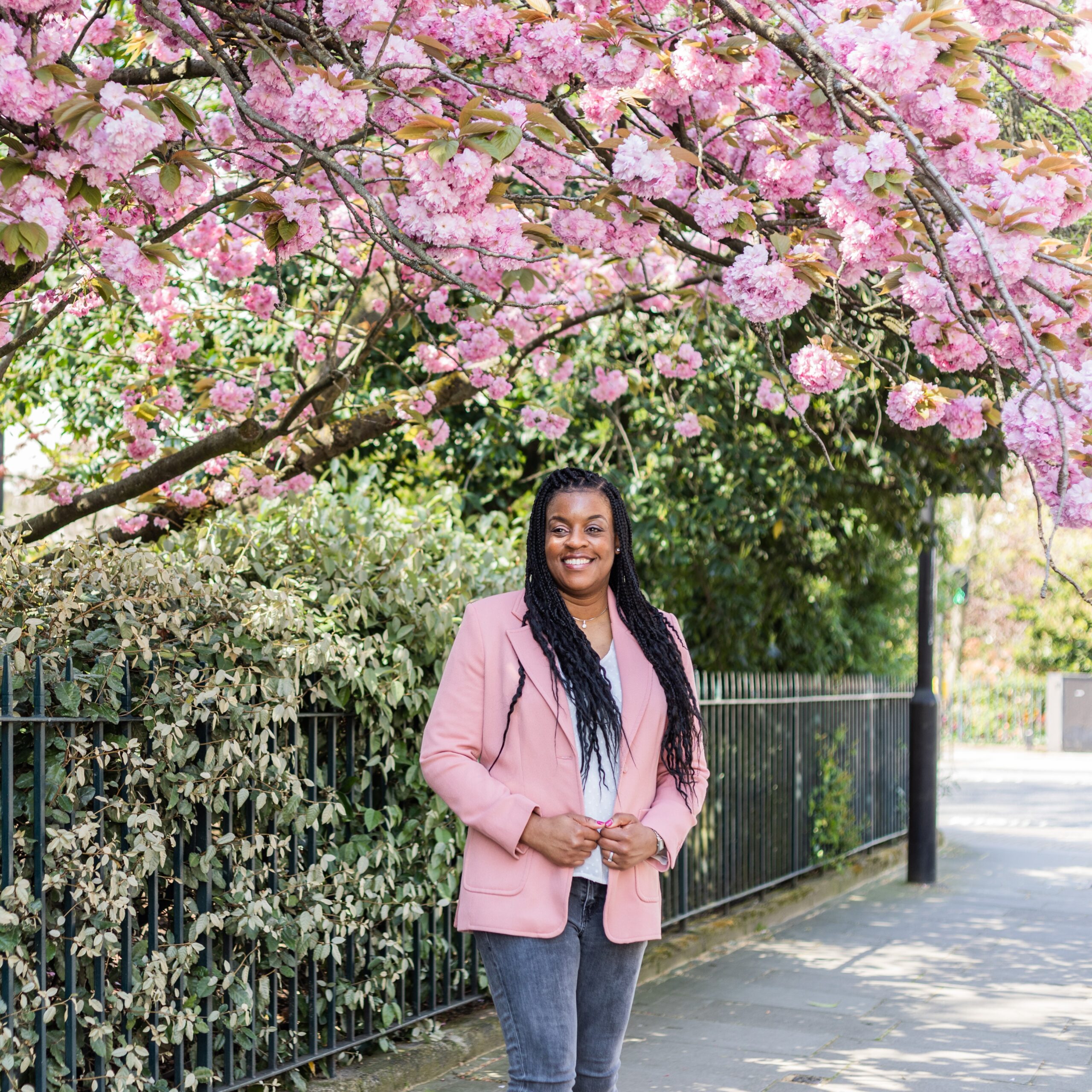 Wedding planner Natasha is stood underneath a blossom tree in London. She is a black lady wearing jeans and a pink blazer. Mini shoot image by London brand photographer AKP Branding Stories