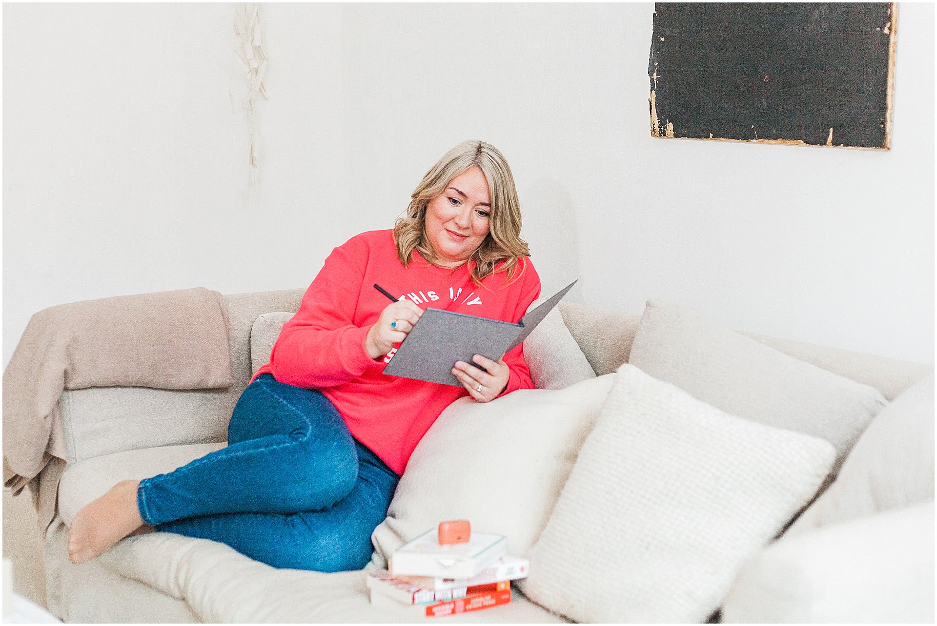 White female with blonde hair in a bright red sweatshirt smiling while writing notes in a journal, comfortably seated on a beige sofa with white and grey cushions, with a black abstract painting on the wall and a stack of books on branding beside her, in a well-lit cosy room.

Image by London brand photographer AKP Branding Stories for the blog post "Brand photography on a budget"