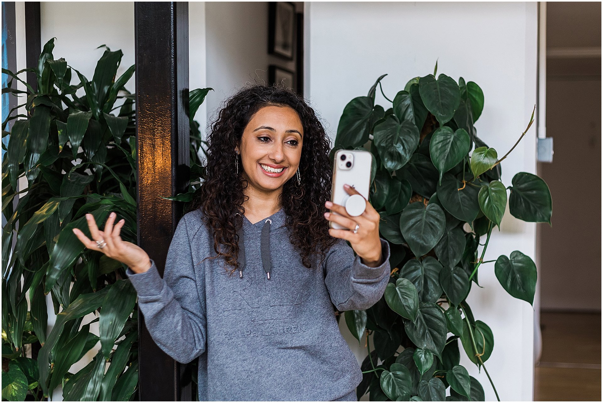 Smiling Asian woman with curly hair taking a selfie, wearing a casual grey hoodie, standing next to a lush green indoor plant, with a black door frame and interior decor in the background

Image by London brand photographer AKP Branding Stories for the blog post "Brand photography on a budget"